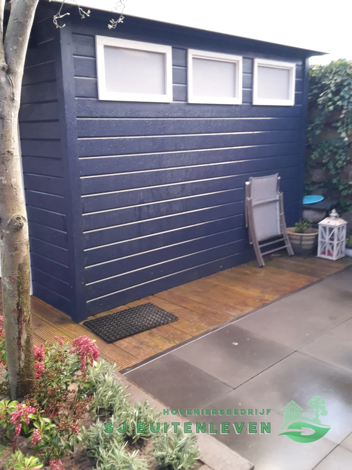 Donkerblauw tuinhuis met drie ramen en houten vlonder, onderdeel van de tuinrenovatie in Hoorn.