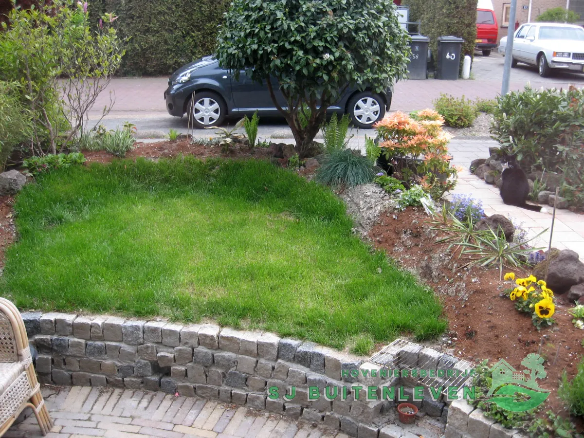 Gazon-eiland in deze voortuin in Middenmeer gezien vanaf de gemetselde kademuur, met een border van vaste planten en sierheesters.