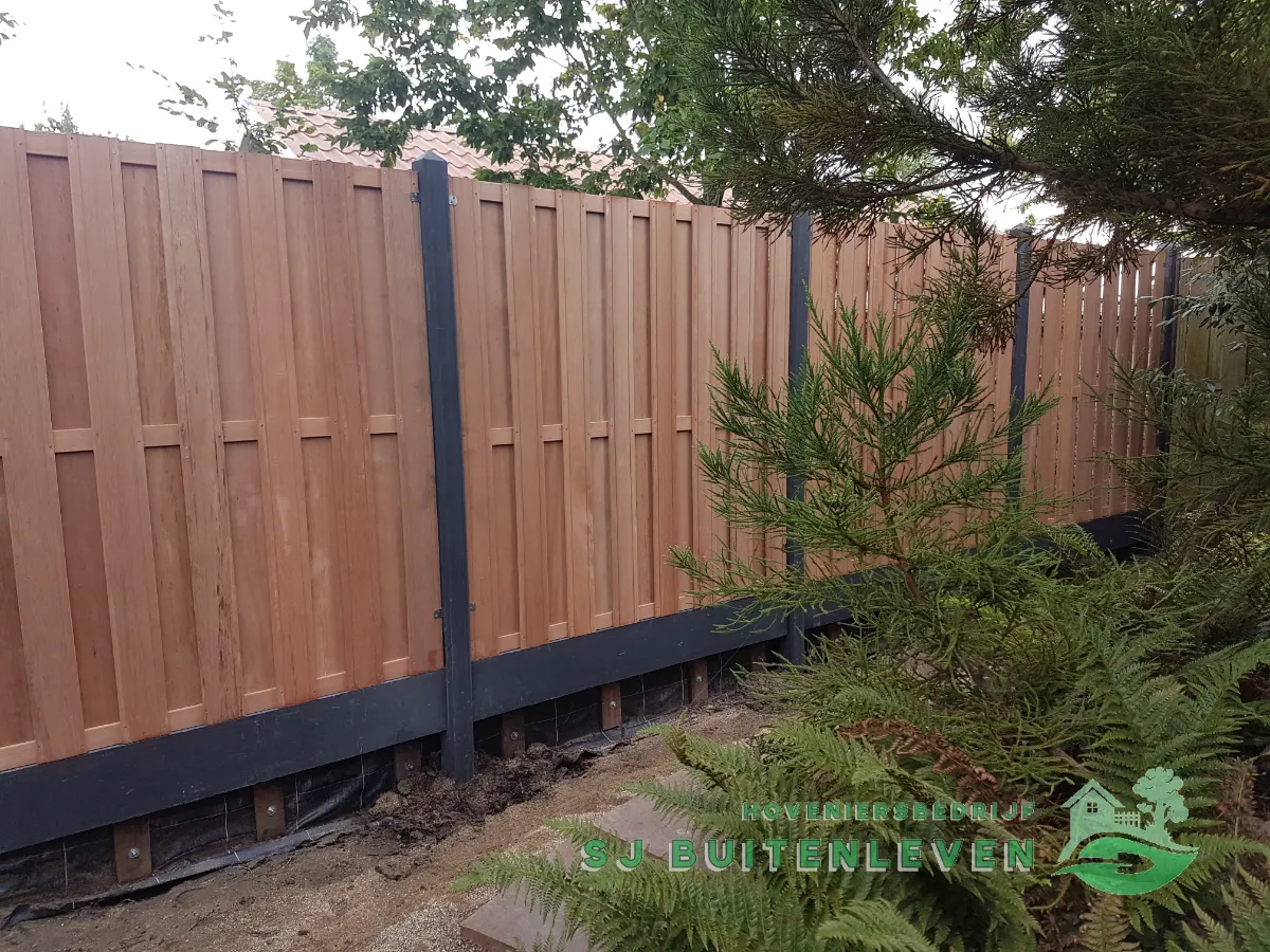Nieuwe hout/betonschutting met verticale planken en zwarte kunststof plint, geplaatst tussen naaldbomen in Nieuwe Niedorp.