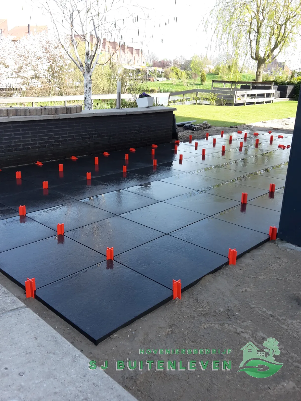 Terrasaanleg in Heerhugowaard met zicht op de vijver, houten vlonder en treurwilg, tegels met kruisjes strak in het verband gelegd tot aan de erfgrens.