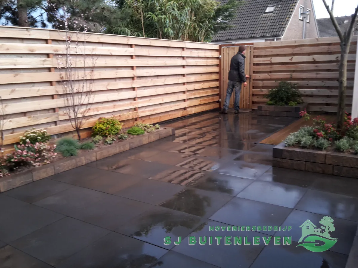 Hovenier loopt door de nieuwe houten poortdeur in de erfafscheiding, met bloeiende border langs de schutting in Hoorn.