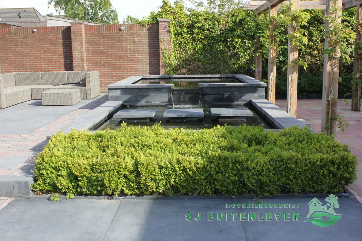 Verhoogde vijver met trapsgewijze waterval, omzoomd door een buxushaag en een loungeset op het afgewerkte terras in Heerhugowaard.