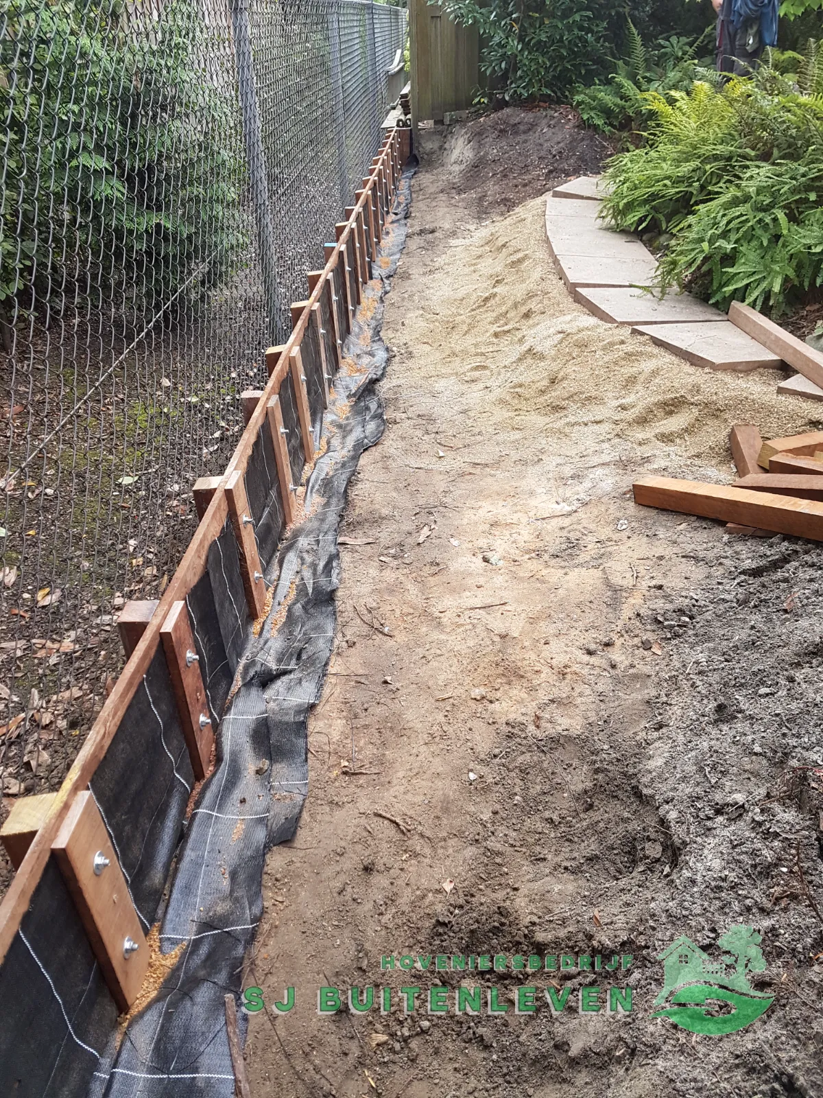 Grondkering in aanbouw met stevige houten palen, zandbed en glooiend tuinpad tussen varens in Nieuwe Niedorp.