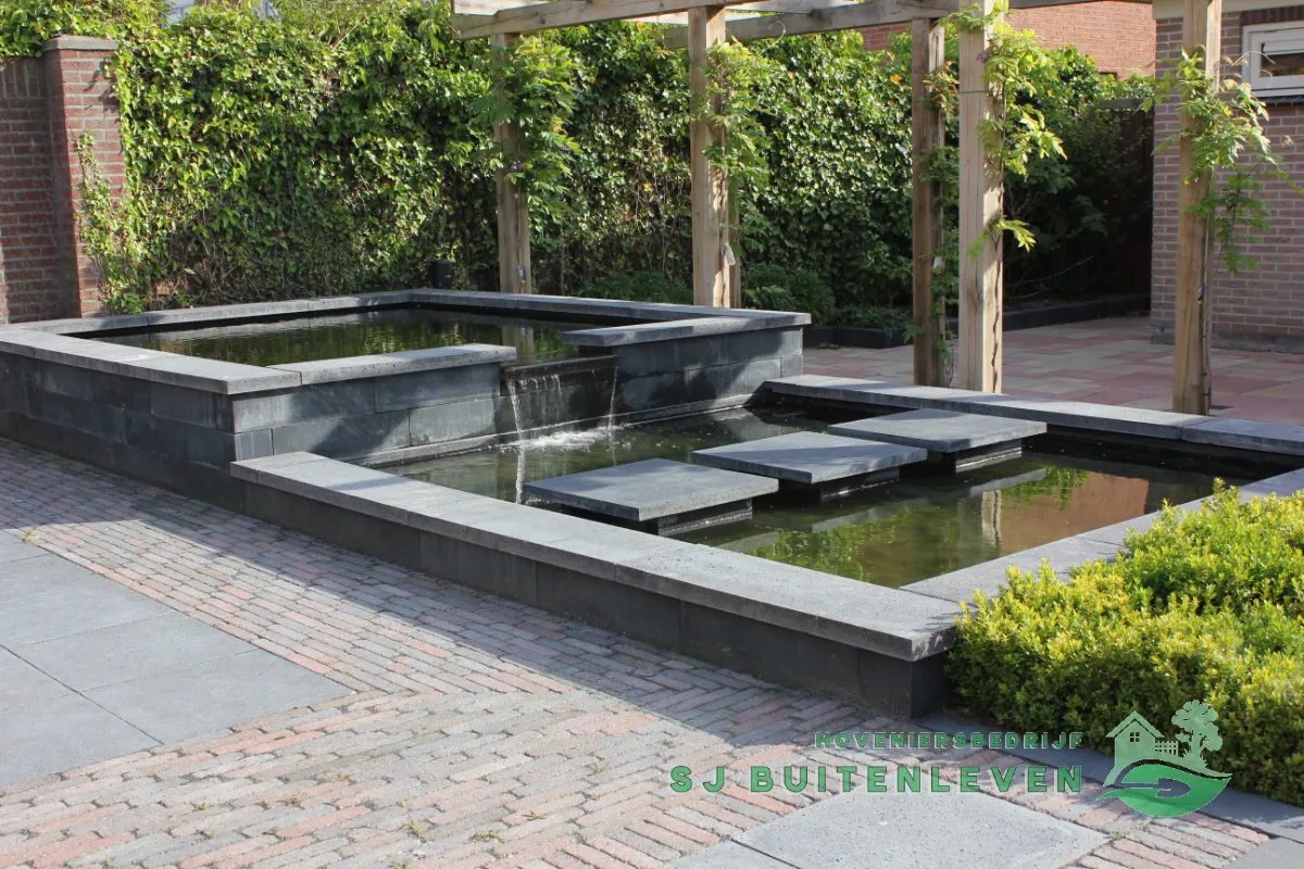 Breder aanzicht van de vijver en pergola, met opstapstenen over het water en een hoge haag als groene achtergrond in Heerhugowaard.