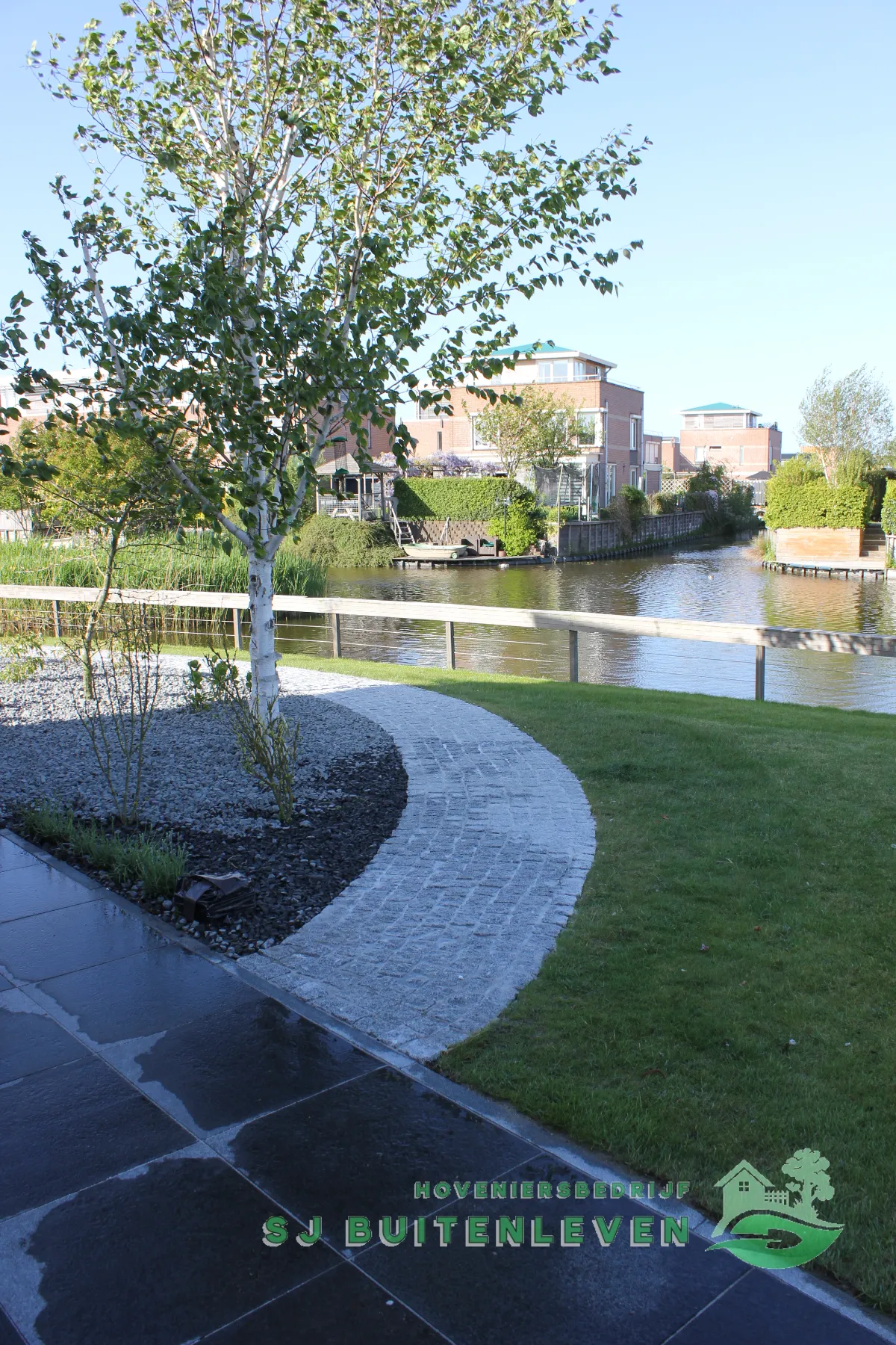 Gebogen pad van natuurstenen kasseien door het gazon in Heerhugowaard, met een border van witte grind rond een berk en een houten hekwerk langs het water.