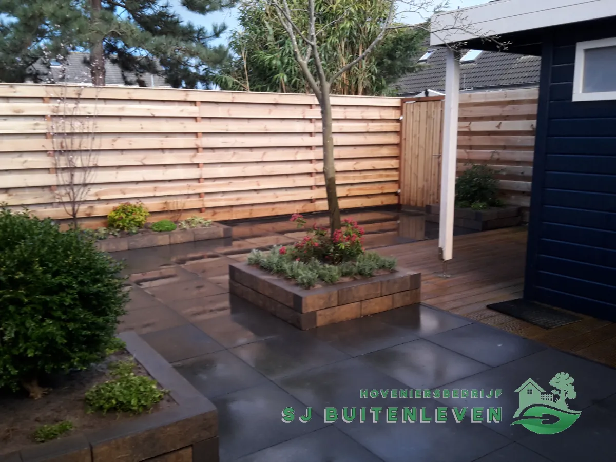 Modern terras met zwarte keramische tegels en verhoogd plantvak met sierboom en rode bloeiende heesters, tuinrenovatie in Hoorn.