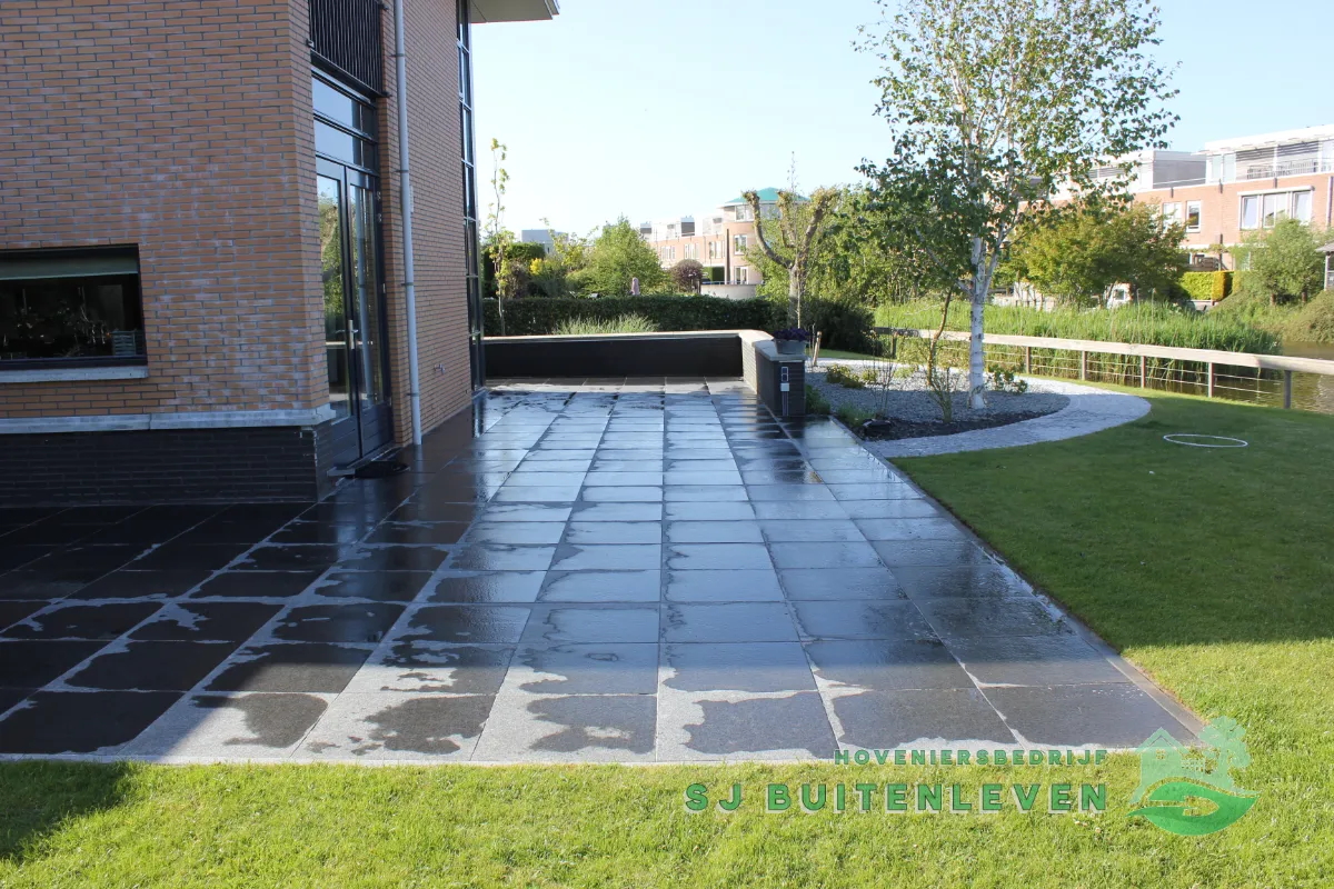 Panoramisch zicht op het afgeronde terras in Heerhugowaard met een border van witte sierkiezels, een jonge berk en de vijver op de achtergrond.