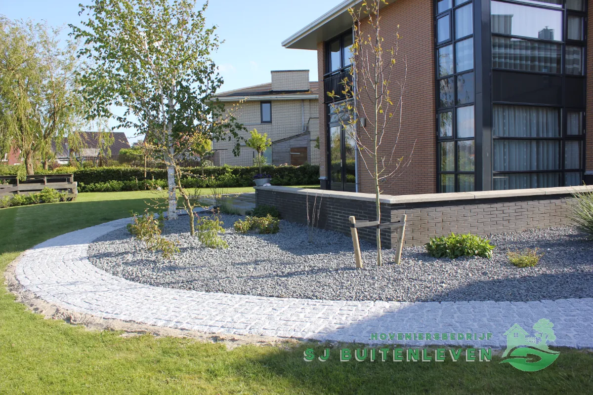 Ronde grindborder met jonge, opgebonden berken en het gebogen kasseipad naast het terras bij de woning in Heerhugowaard.