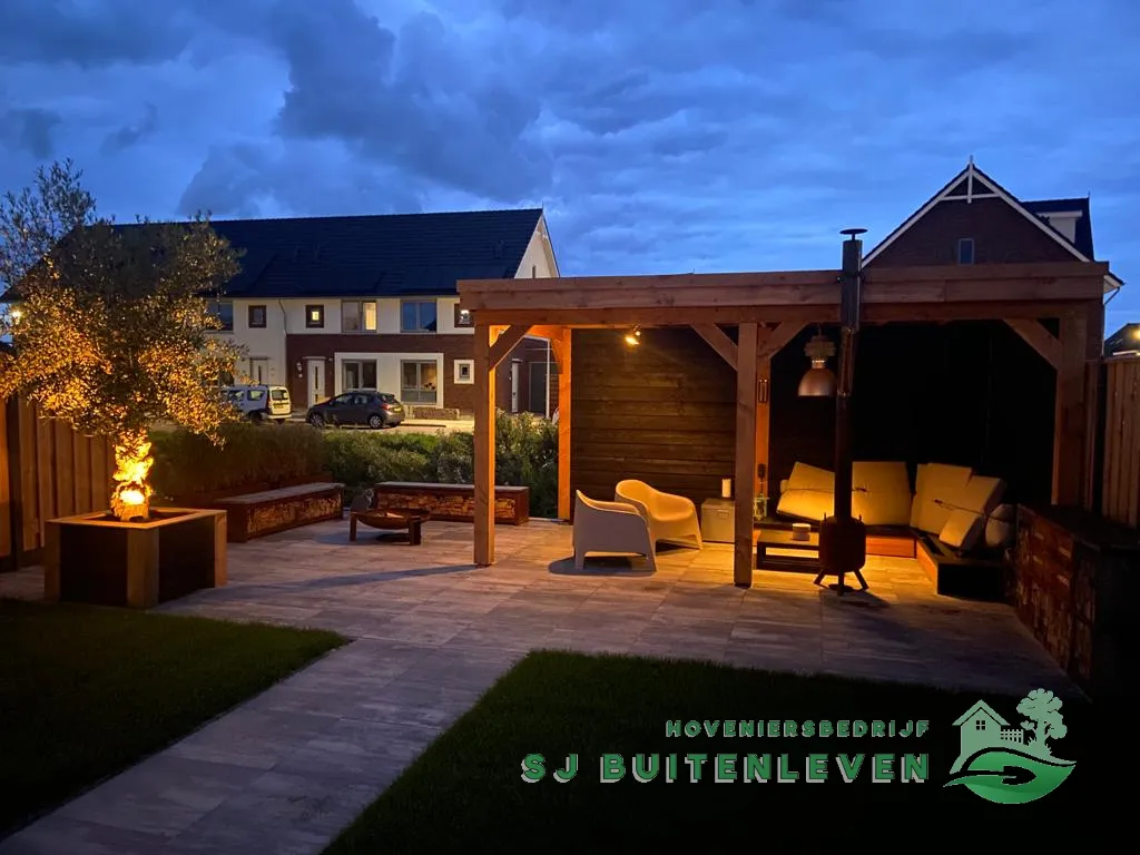 Sfeervolle avondfoto van de afgeronde tuin in Spanbroek met verlichte pergola, loungebank en brandende vuurschaal.