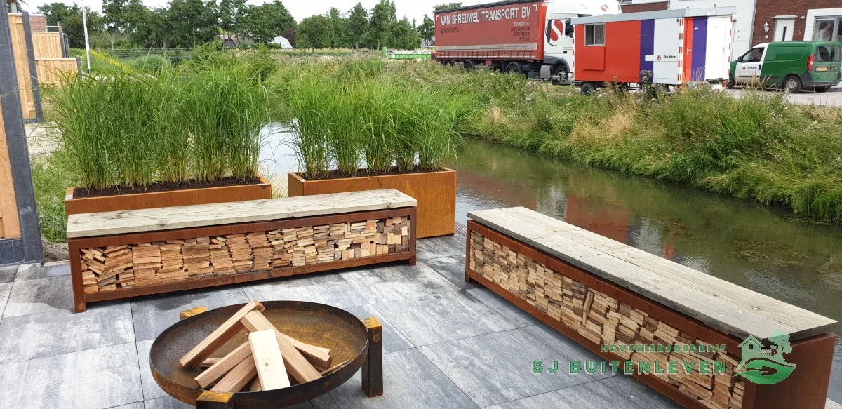 Tuinaanleg in Spanbroek: afgeronde zitplek met cortenstalen vuurschaal en houtopslagbanken aan de waterkant.
