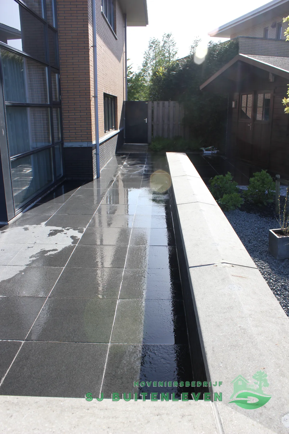 Zijpad met terrastegels langs de woning in Heerhugowaard, leidend naar een houten poort met een tuinhuisje en heg van de buren op de achtergrond.